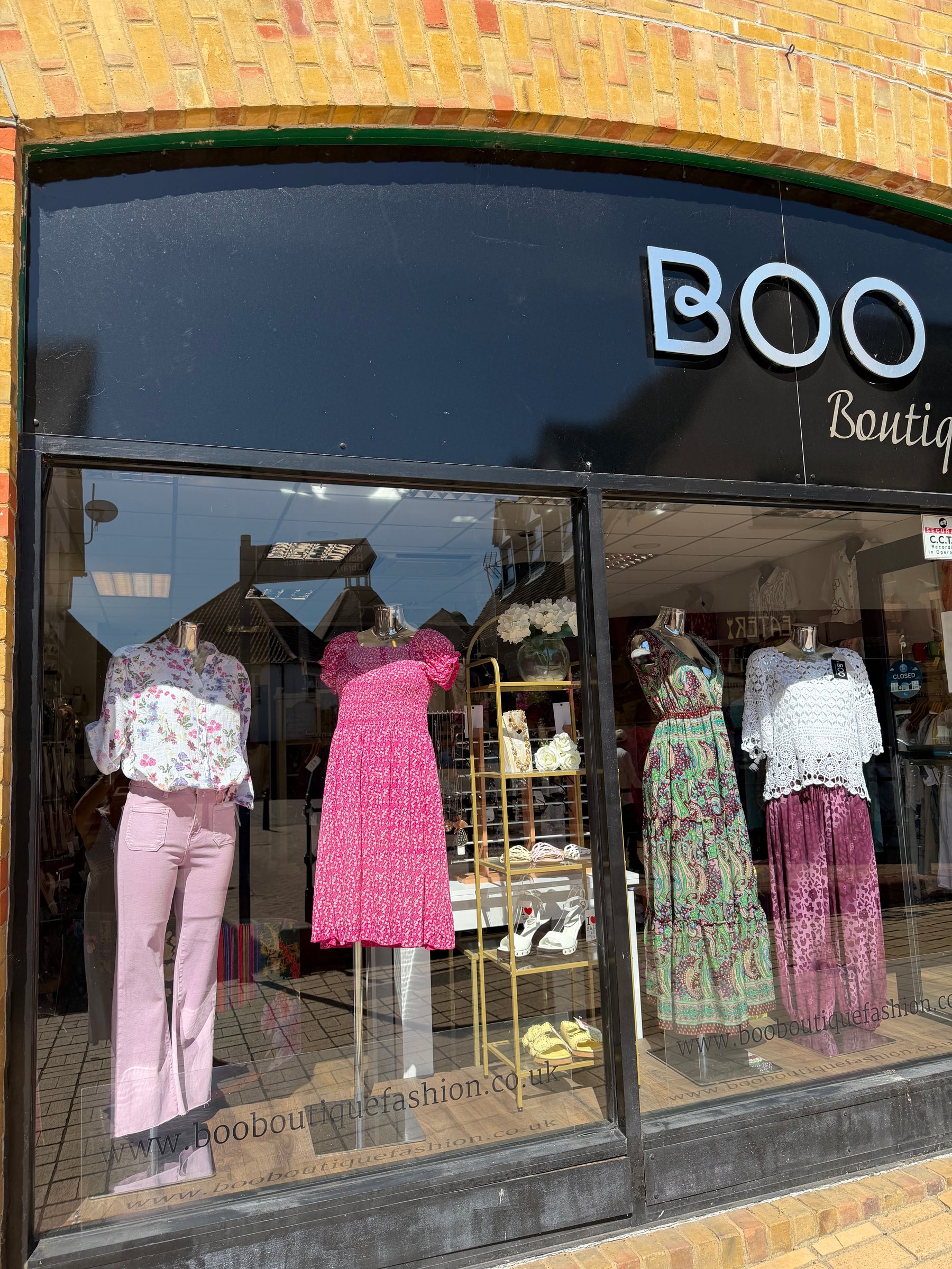 A storefront window display for BOO Boutique, showing several mannequins wearing colorful outfits including a pink dress, patterned top and trousers, and a white lace top with patterned pants. The shop has a black arched sign area with the BOO Boutique name and a brick facade above.